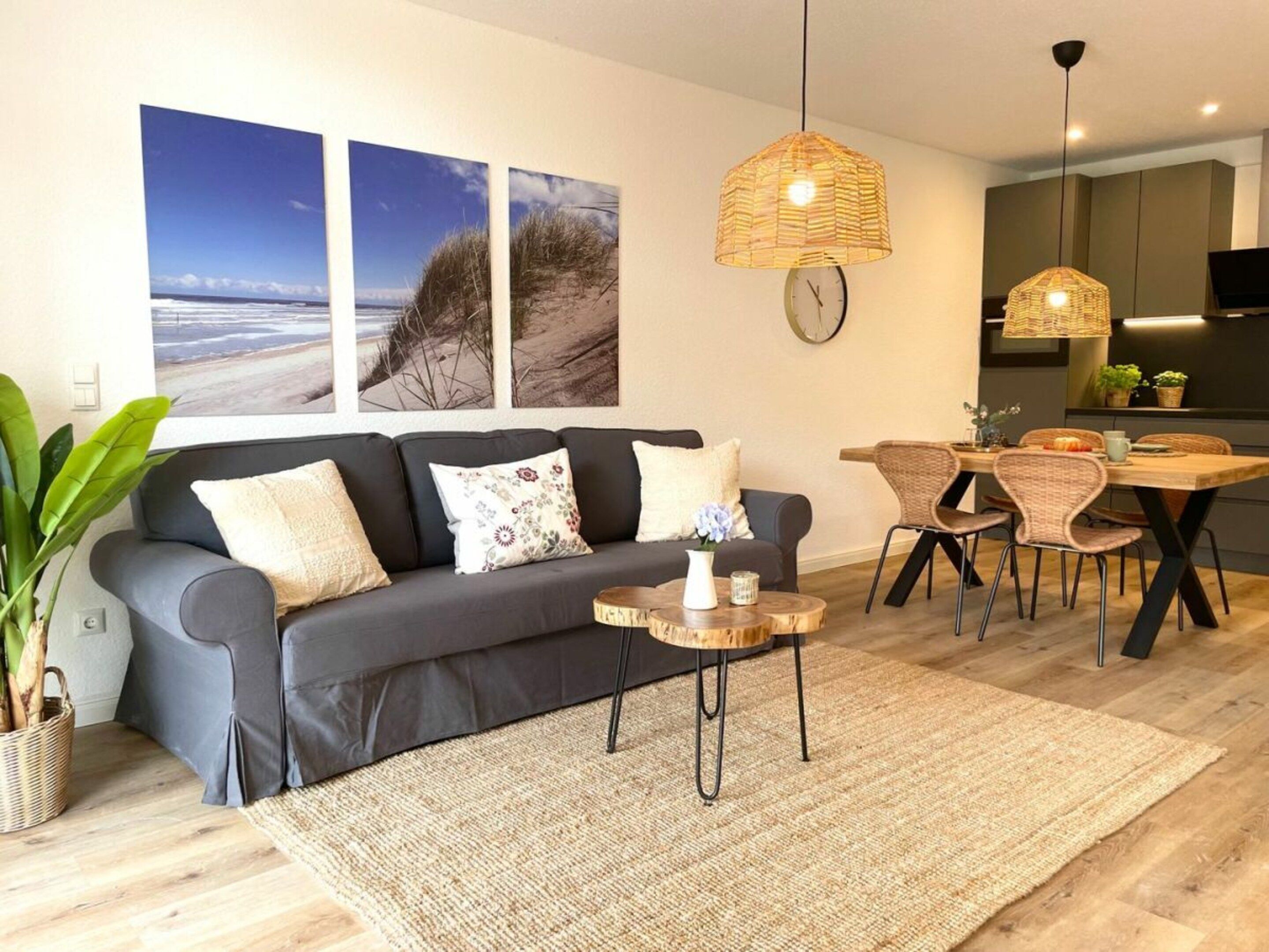 Una sala de estar moderna con un sofá gris, una mesa de comedor con sillas de mimbre y una planta grande.