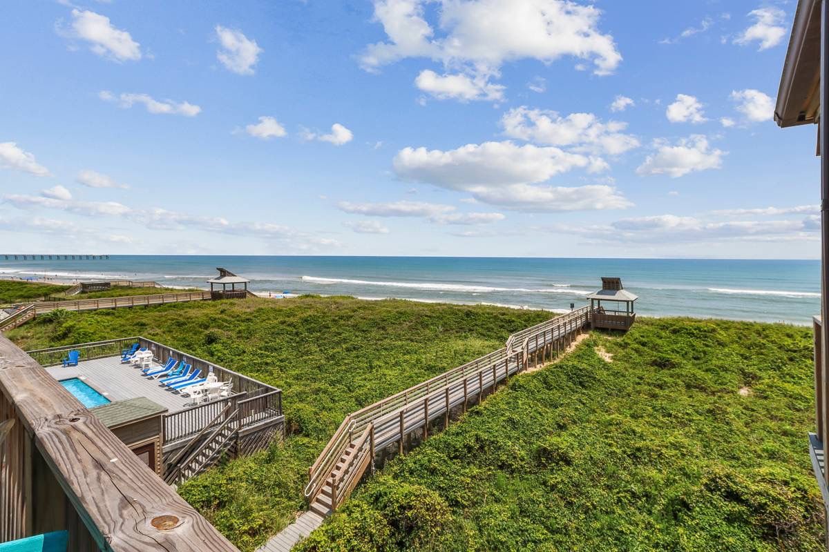 A wooden deck with a pool and lounge chairs overlooking a beach and ocean.