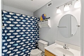 Ein Badezimmer mit Duschvorhang mit Fischmuster, Toilette, Waschbecken mit Spiegel und Ablagefläche für Toilettenartikel.