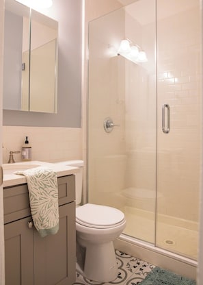 A bathroom with a toilet, a sink with a towel, a mirror, and a shower enclosure. - Classic NYC 3BR/1BA City Escape! Stay Near Central Park! (New York)