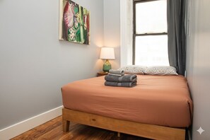 A neatly made bed with a brown bedspread and folded towels, a green lamp on a bedside table, and a colorful abstract painting on the wall. - Classic NYC 3BR/1BA City Escape! Stay Near Central Park! (New York)