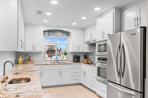 A modern kitchen with white cabinets, a stainless steel refrigerator, and a window with a view of the outside. - Hook, Wine and Sinker ~3 Bed / 2 Bath Home with a Pool! (Rockport)