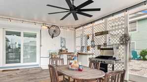 Eine Terrasse im Freien mit Holztisch und -stühlen, einem Deckenventilator, Lichterketten und einem Grill.