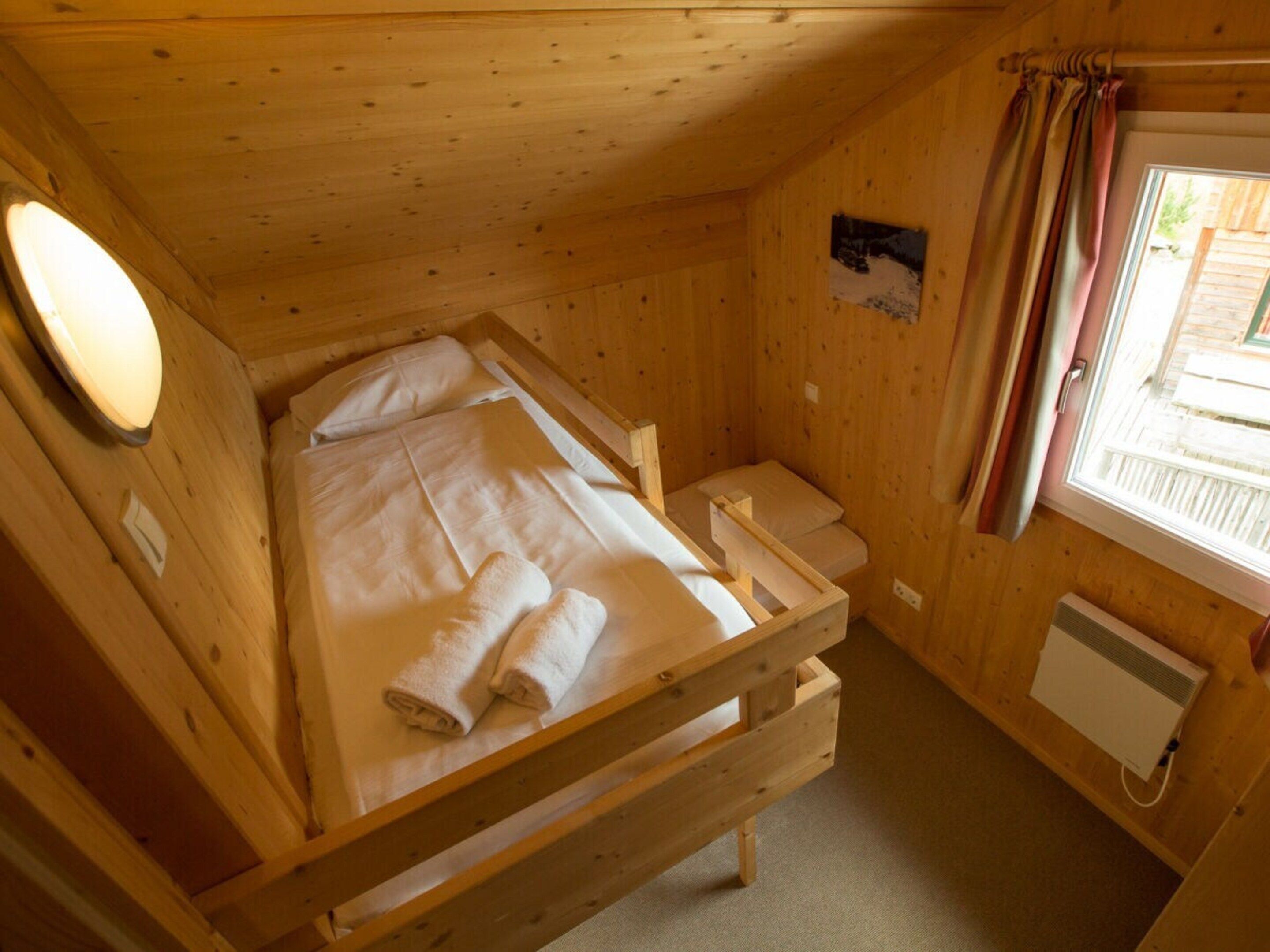 Eine kleine Holzhütte mit einem Einzelbett, einem Fenster mit Vorhängen und einem Heizkörper an der Wand.