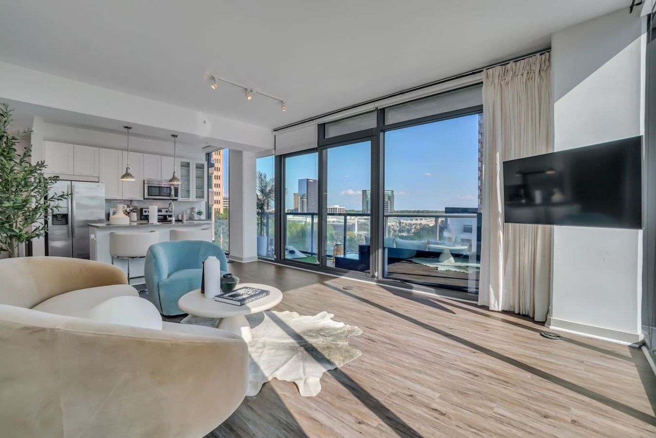 Una sala de estar moderna con sofá, sillón, mesa de centro, ventanas amplias con vista a la ciudad y un televisor de pantalla plana montado en la pared.