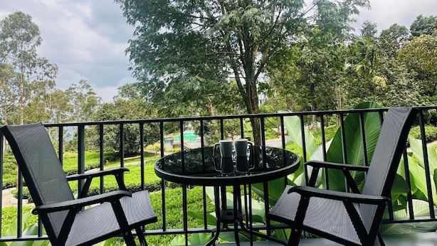 A balcony with a round table and two chairs, surrounded by a metal railing, overlooking a garden with trees and a clear sky. - WAYANAD DE MOUNT RESORT BY RAK ROOMS (KL)