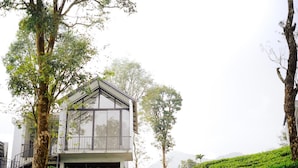 A modern building with a glass facade, surrounded by lush greenery and a white wall. - WAYANAD DE MOUNT RESORT BY RAK ROOMS (KL)