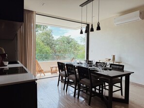 Een eethoek met een tafel voor vier personen, een keuken met een fornuis en een groot raam met uitzicht op bomen.