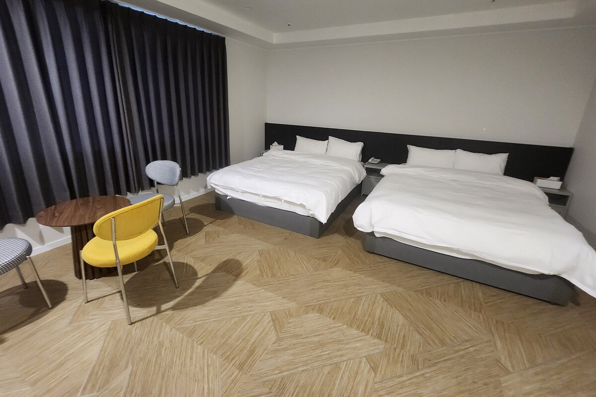 Hotellihuone, jossa on kaksi sänkyä, pieni pyöreä pöytä, keltainen tuoli ja suuri ikkuna, jossa on tummat verhot.