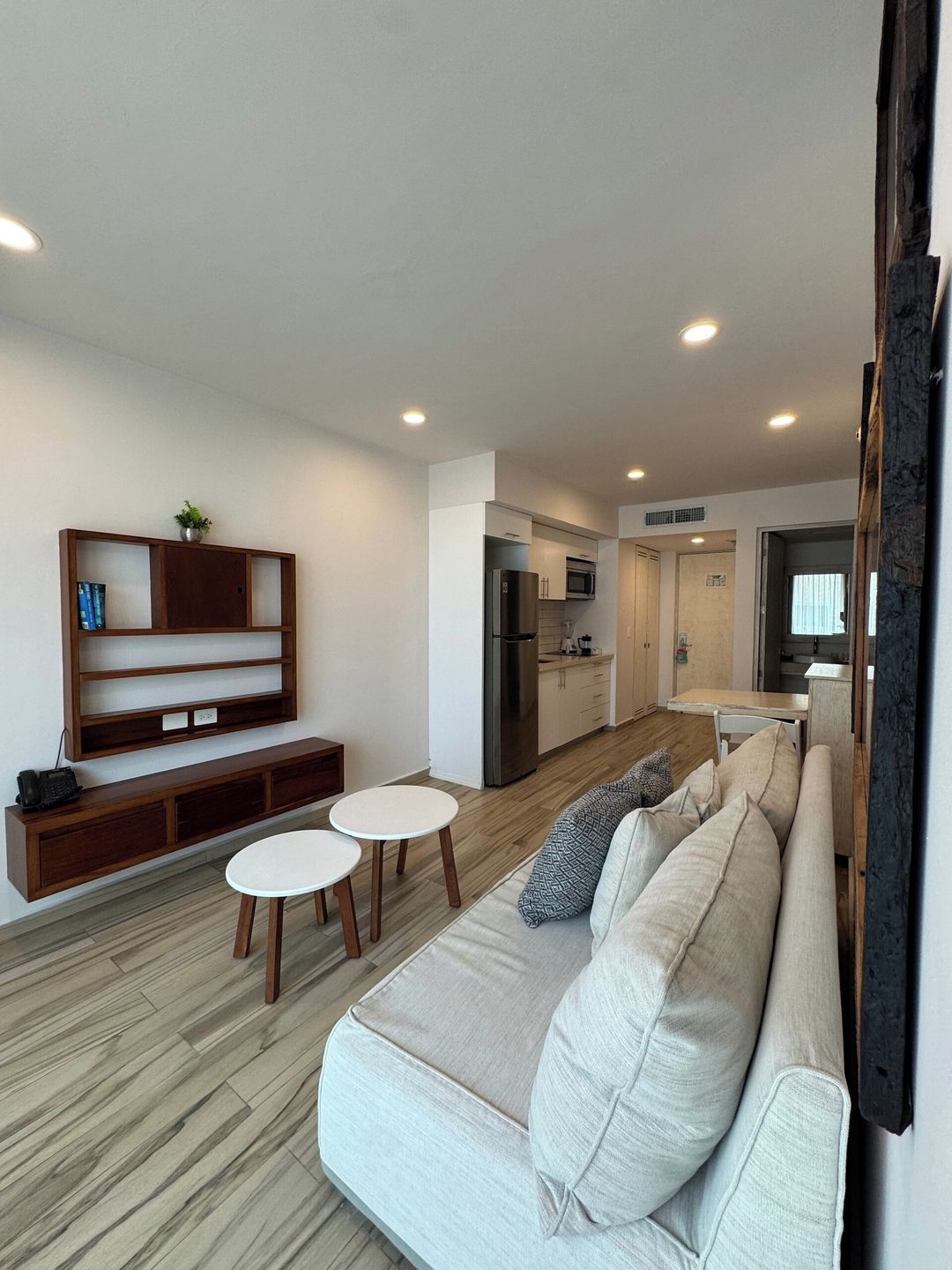 A modern living room with a sofa, two small tables, a wooden console, and a kitchen in the background.