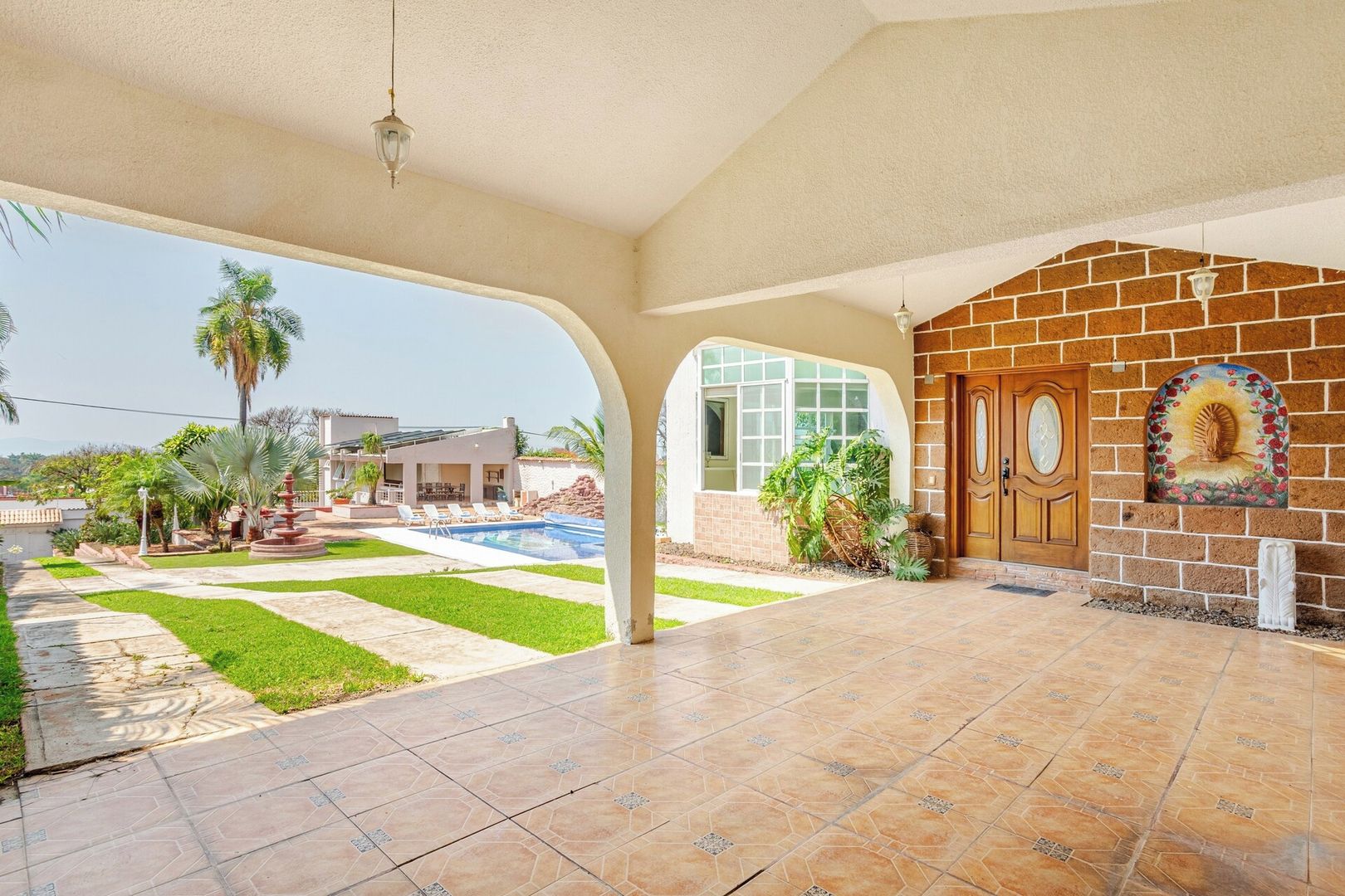 A covered patio area with a swimming pool, a brick wall with a decorative element, and a view of a residential building in the background.