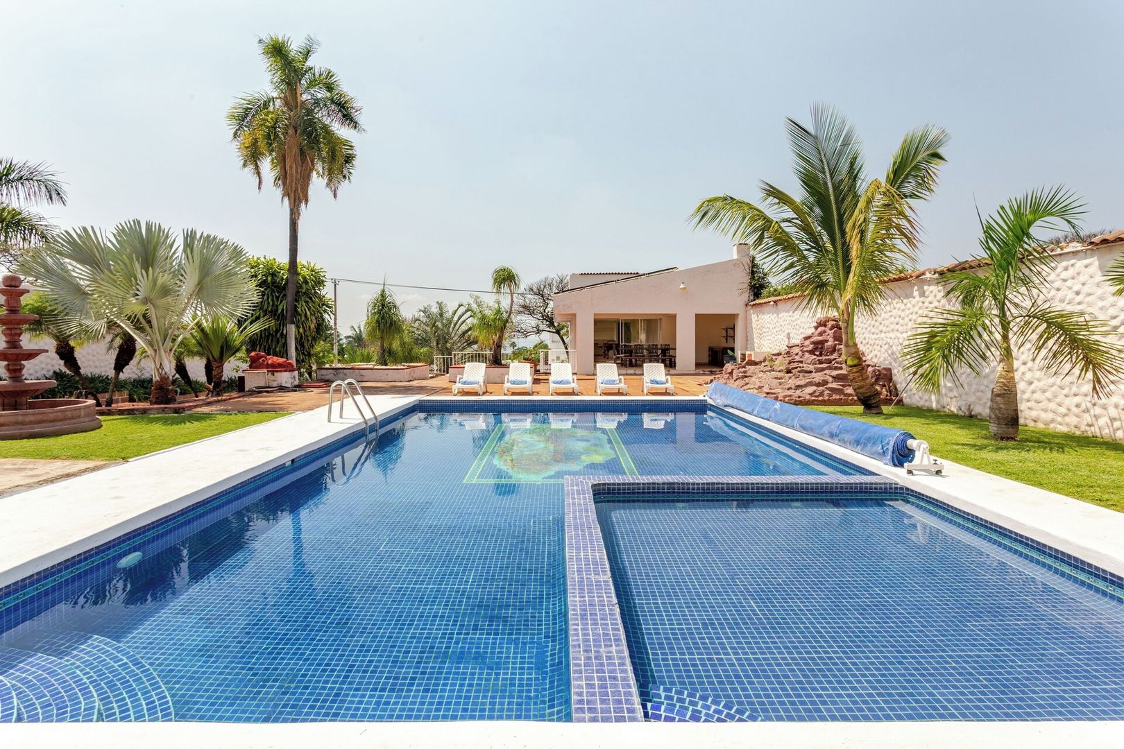 A rectangular swimming pool with blue tiles, surrounded by white lounge chairs, palm trees, and a building with a tiled roof.