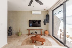 A modern living room with a flat-screen TV, a wooden bench, a woven rug, and a stone wall. - Aldea Zama · 2BR with pool · Guest favorite (Tulum)