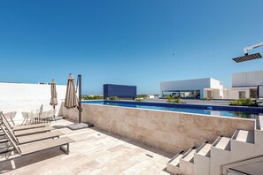 A rooftop pool area with a clear view of the sky, a modern building, and a lounge with chairs and umbrellas. - Aldea Zama · 2BR with pool · Guest favorite (Tulum)