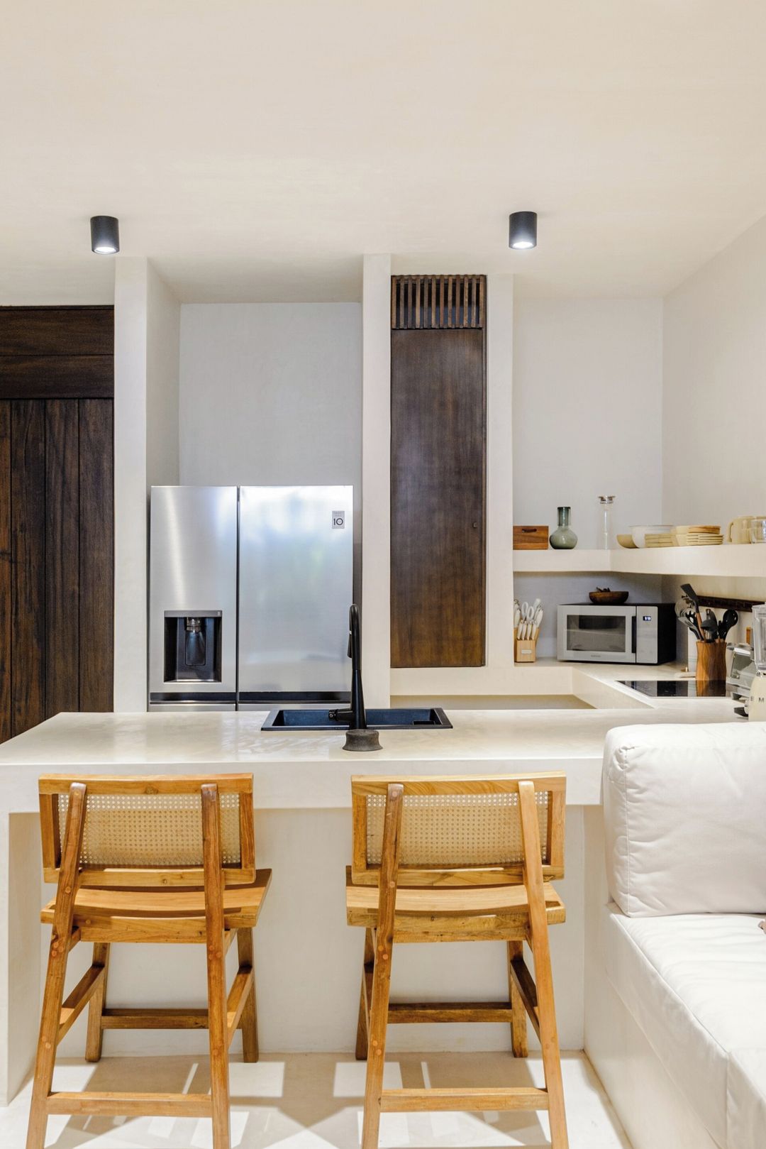 A modern kitchen with a central island, two wooden chairs, a white sofa, and a dark wooden accent wall.