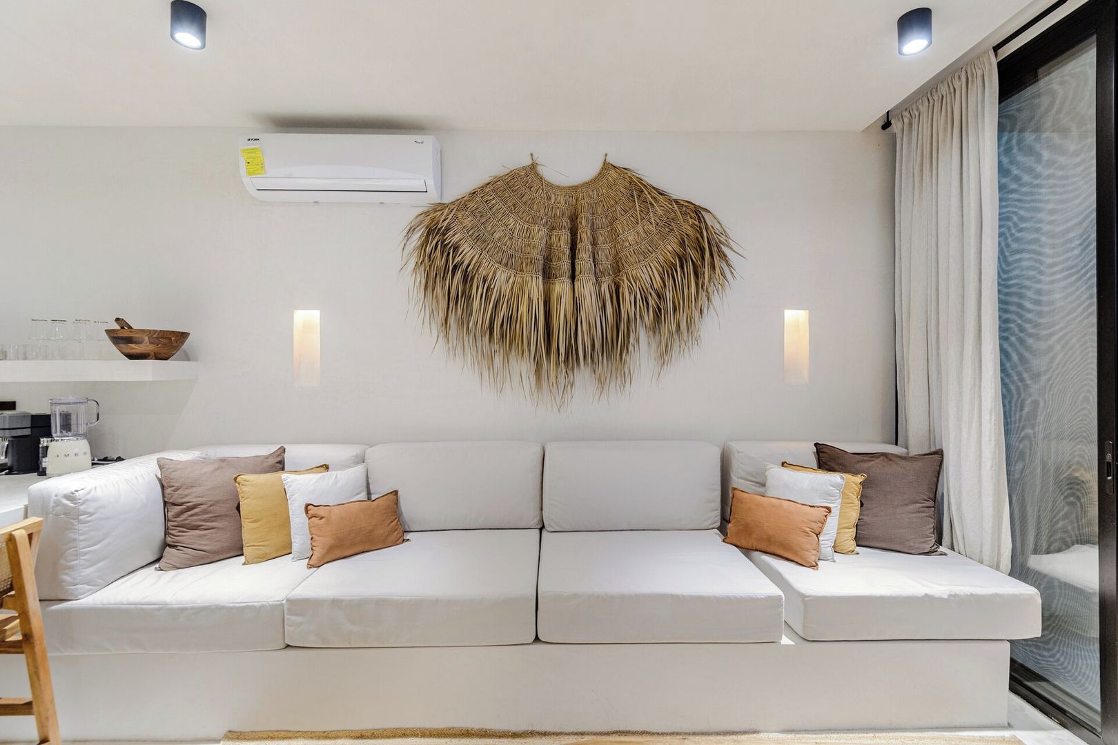 A modern living room with a white sofa, decorative wall hanging, and a glass door with curtains.
