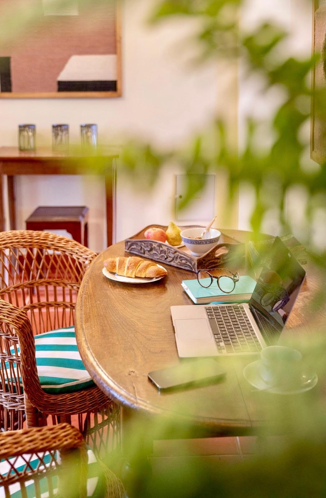 Ein runder Holztisch mit einem Laptop, einer Brille, einer Schüssel mit einem Löffel, einem Teller mit einem Croissant und einem Buch.