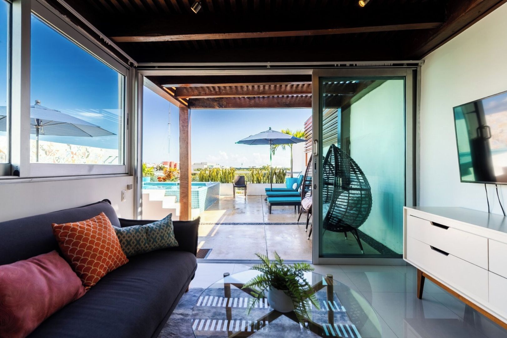 Ein modernes Wohnzimmer mit einer Couch, einem Glastisch, einem Fernseher und Blick auf eine Außenpatio mit Pool und Sitzgelegenheit.