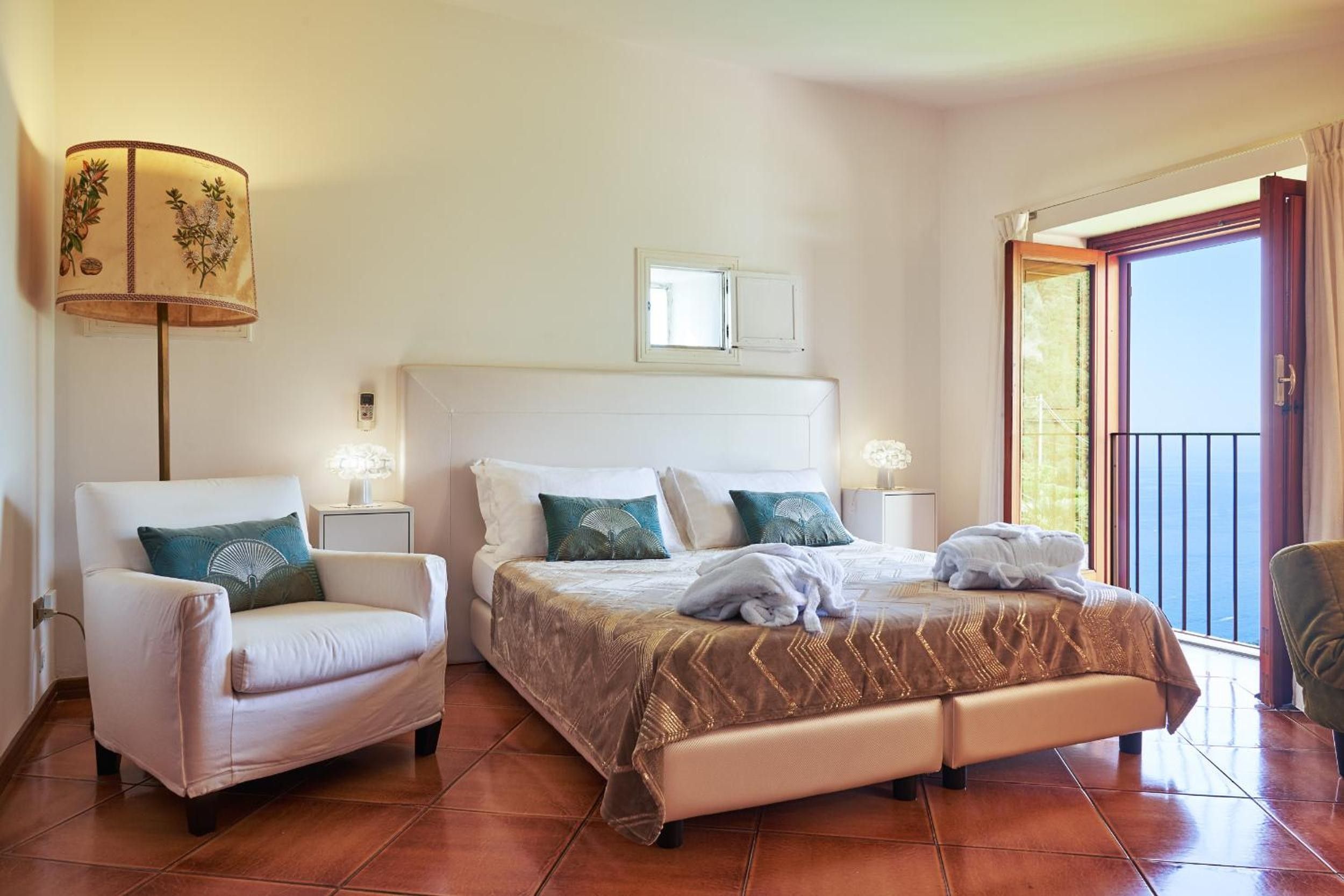 Habitación de hotel con cama, dos mesitas de noche, una silla y una lámpara, con vista al exterior.