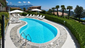 Piscina curva com espreguiçadeiras e guarda-sóis, em meio a azulejos de pedra e vegetação, com vista para montanhas e céu claro.