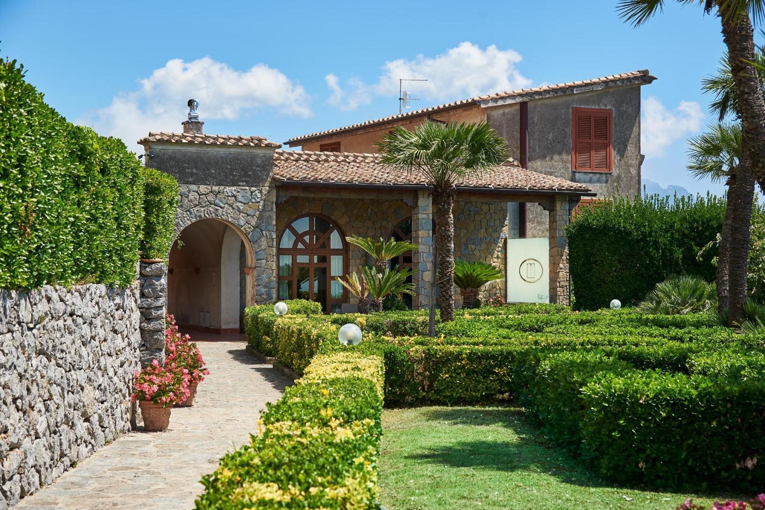 Een gebouw in traditionele Mediterrane stijl, met een gevel van natuursteen, een pannendak en een verzorgde tuin met hagen en palmbomen.
