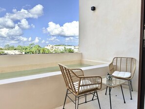 A balcony with two wicker chairs and a small table, a pool, and a view of buildings and trees under a partly cloudy sky. - Please note: electricity is not included in reservations longer than 28... (Tulum)