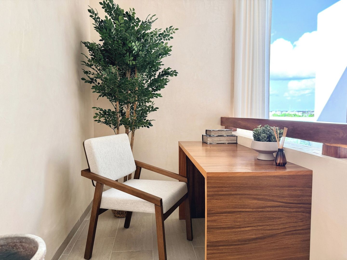 A corner of a room with a wooden desk, a chair, a potted plant, and a window with a view.