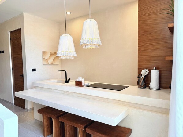 A modern kitchen with a white countertop, two pendant lights, a built-in stove, and wooden stools. - Please note: electricity is not included in reservations longer than 28... (Tulum)