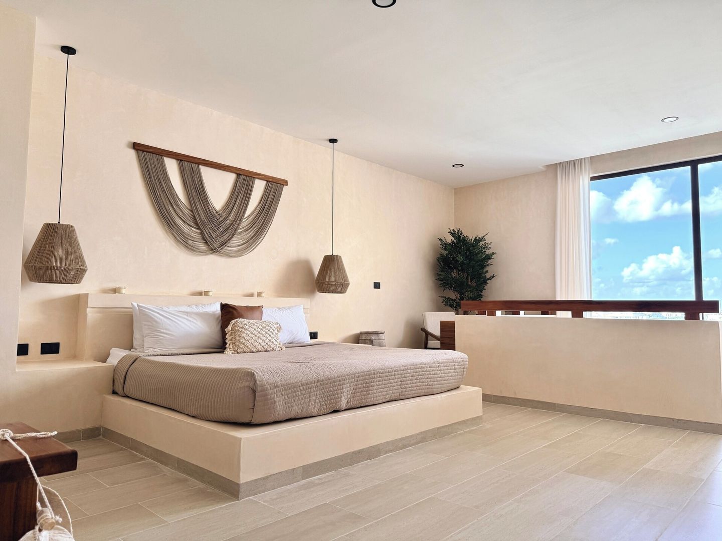 A modern bedroom with a large bed, pendant lights, a wall-mounted macrame piece, and a view of the sky through a window.