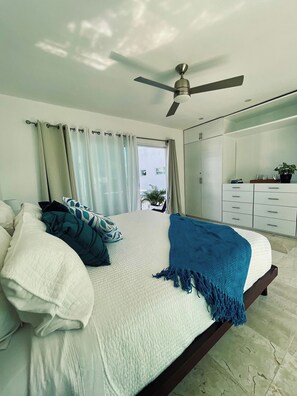 Ein Schlafzimmer mit Bett, Vorhängen, Deckenventilator, Blick durch ein Fenster und einem Badezimmer mit Spiegel und Schränken.