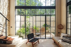 A spacious room with a large window, a sofa, a chair, and a staircase leading to an outdoor area with plants. - Townhouse with private pool facing the jungle (Tulum)