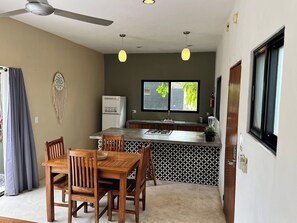 Eine kompakte Küche mit Kühlschrank, ein Essbereich mit Tisch und Stühlen, Pendelleuchten und ein Fenster mit Blick auf Grünflächen.