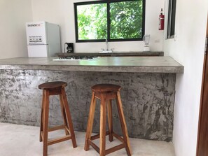 Eine Küche mit Betonarbeitsplatte, zwei Holzschemeln, einem Gasherd und einem Fenster mit Blick auf Bäume.