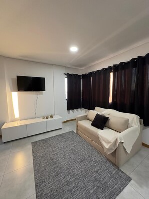 A modern living room with a flat-screen TV, a white entertainment unit, a beige sofa, and a grey rug. - Aconchegante, moderno e completo URU 602 (Joinville)