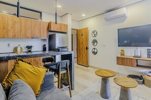 A modern kitchen with wooden cabinets, a marble countertop, and a built-in stove. There is a dining area with a gray sofa, a yellow throw pillow, and two round tables. - Charm and comfort apartment in Aldea Zama (Tulum)