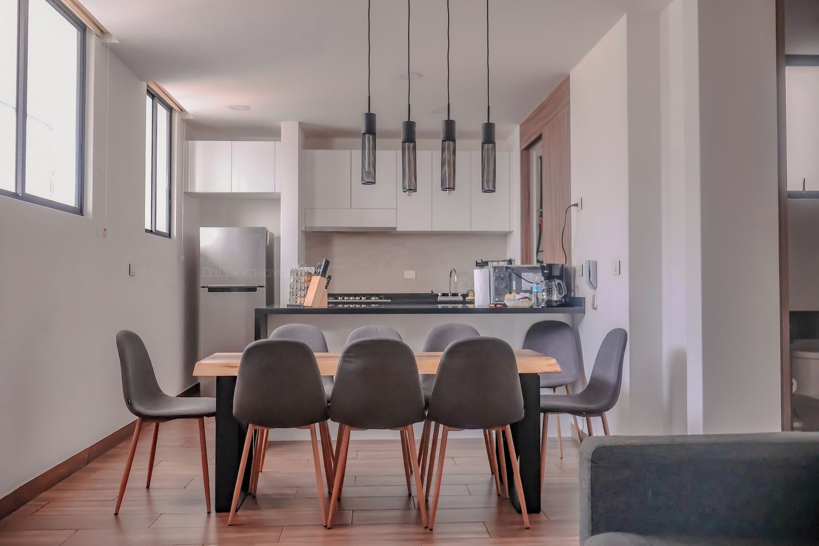 A modern kitchen with a dining table and chairs, a refrigerator, and hanging pendant lights.