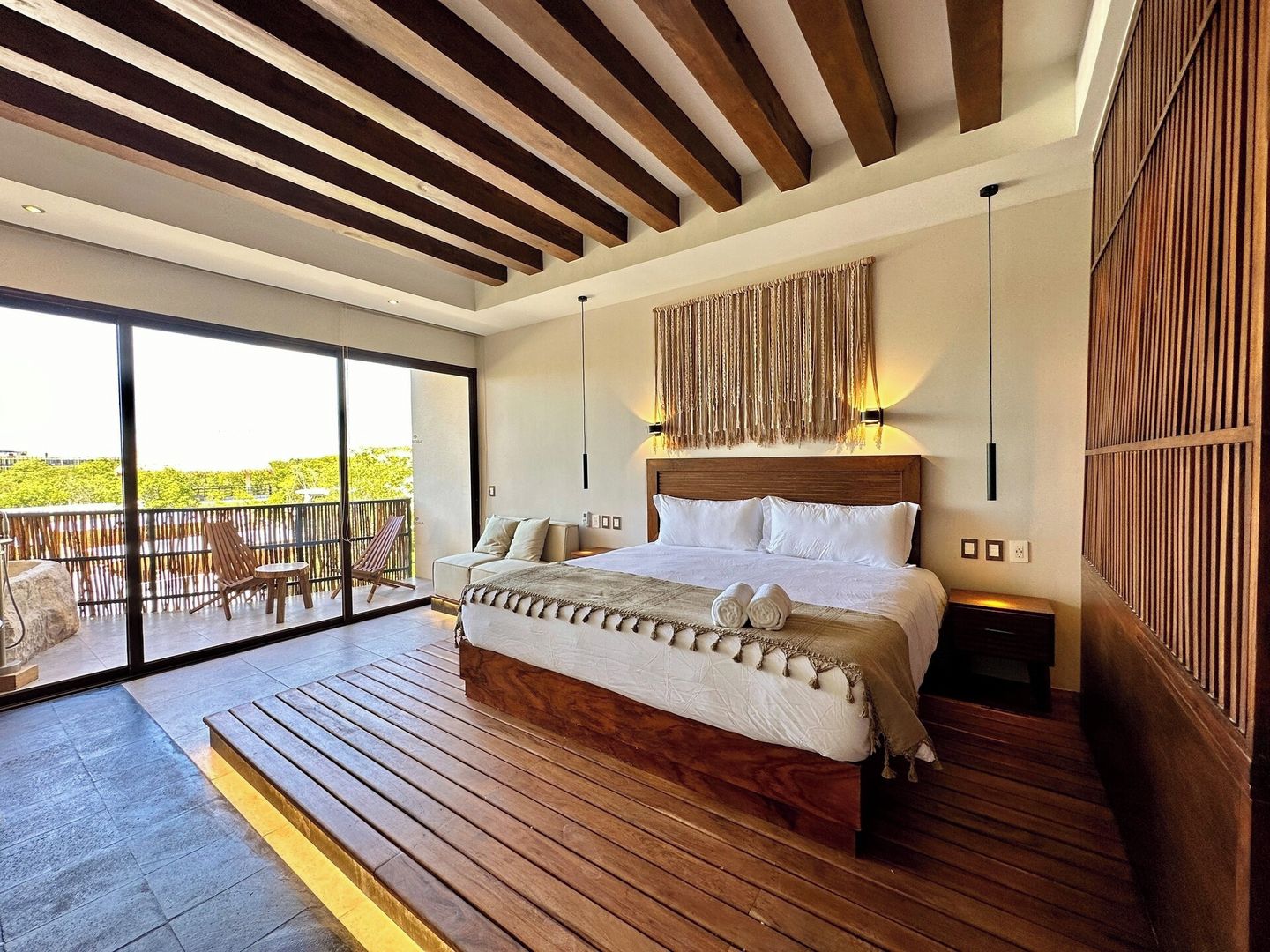 A modern bedroom with a large bed, a balcony with outdoor furniture, and a wooden ceiling.