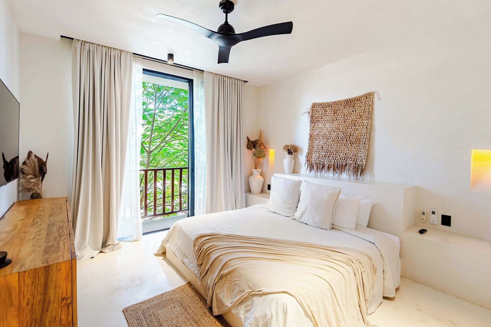 Una camera da letto con un letto grande, un ventilatore a soffitto, un balcone con tende e un lampadario a parete.