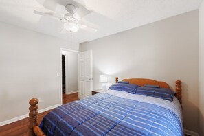 Ein Schlafzimmer mit einem Bett, einem hölzernen Kopfende, einem Deckenventilator, einem Nachttisch und einer Tür zu einem anderen Raum.