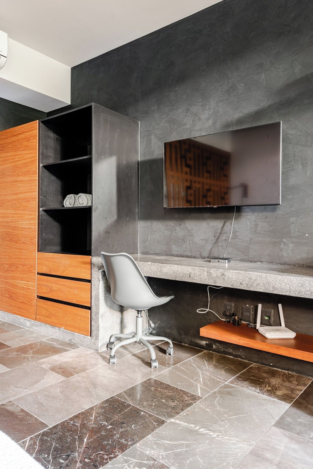 A modern office space with a built-in desk, a chair, a flat-screen TV mounted on the wall, and a shelf with rolled-up documents.
