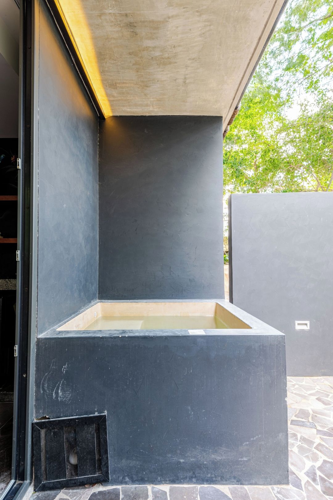 A modern outdoor bathtub area with a concrete structure, a small window, and a stone-tiled floor.