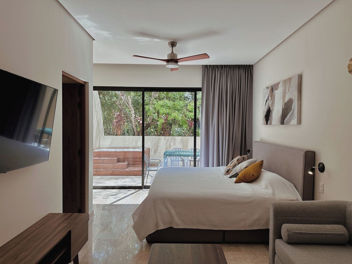 A bedroom with a bed, a TV, a ceiling fan, a window with curtains, and a view of the outdoors.