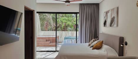 A bedroom with a bed, a TV, a ceiling fan, a window with curtains, and a view of the outdoors.