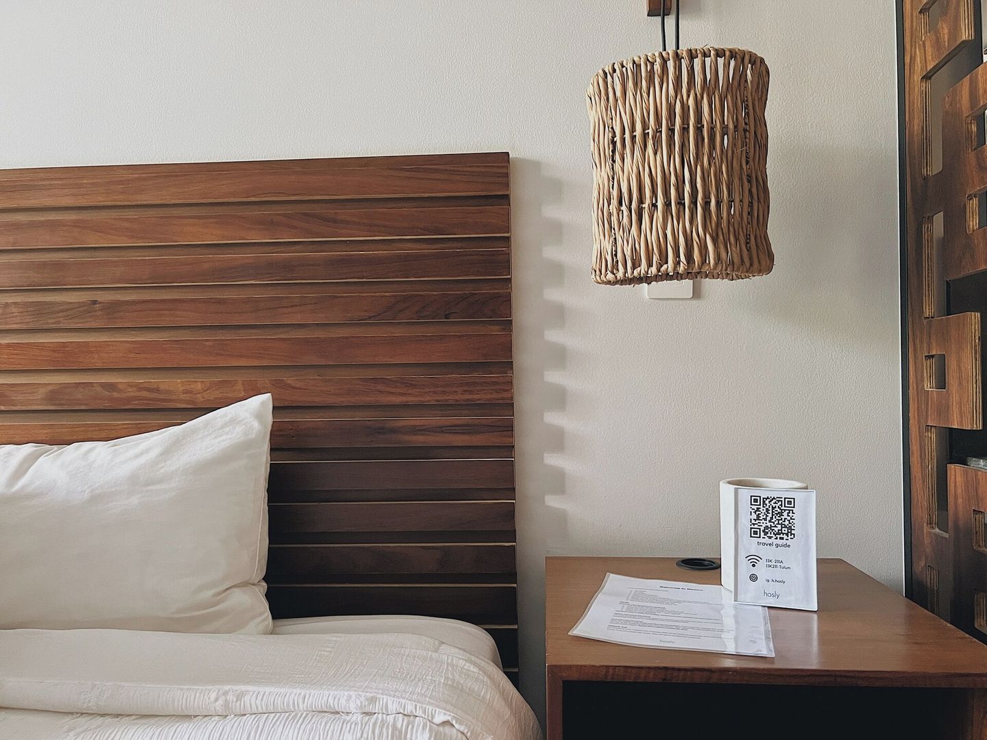 Una camera d'albergo con una testiera in legno, un lampadario intrecciato, un comodino con una guida turistica e una porta con pannelli in legno.
