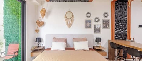A bedroom with a large bed, two bedside tables, a desk with chairs, a green wall, and a view of a green outdoor area.