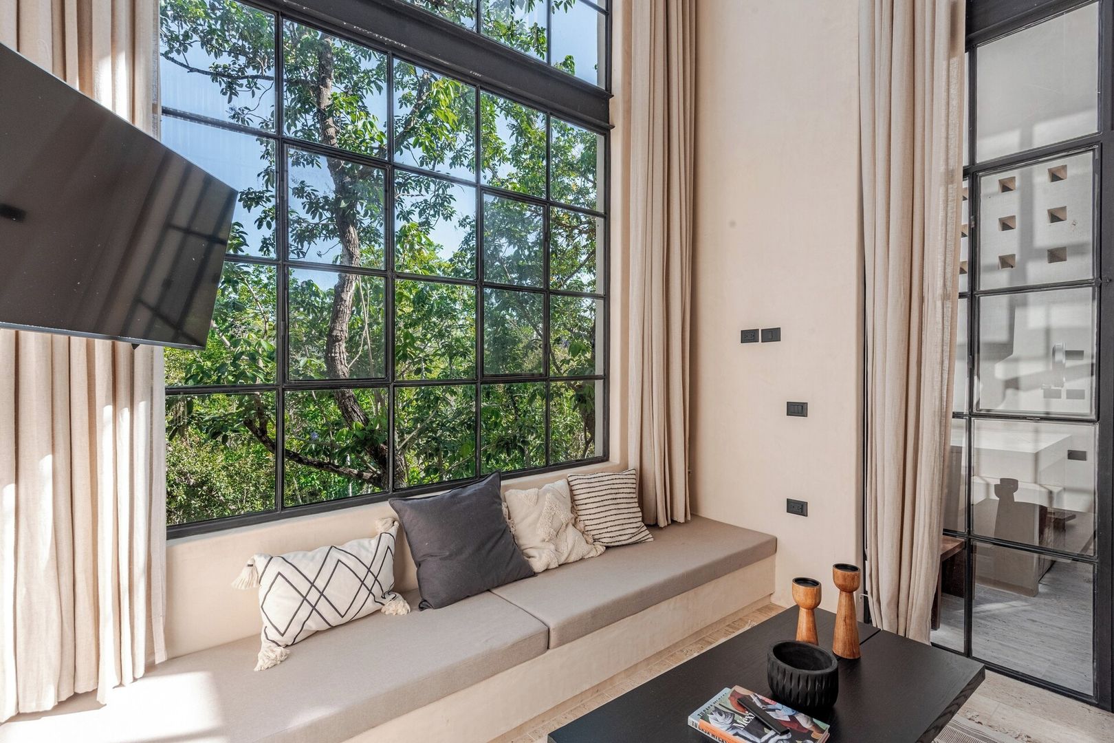 A room with a large window, a sofa with cushions, a table with a magazine, and a lamp.