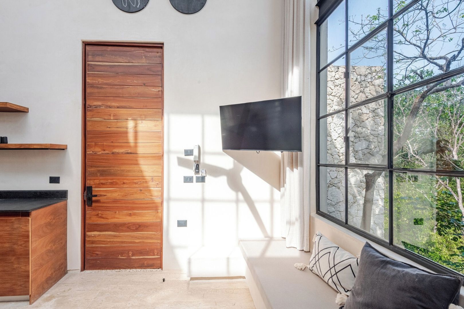 Una camera moderna con una porta in legno, una TV a schermo piatto fissata al muro e un'ampia finestra con vista sugli alberi.