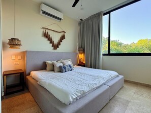 Ein Schlafzimmer mit einem großen Bett, einer Klimaanlage, einer Wandleuchte und Blick auf Bäume durch ein Fenster.