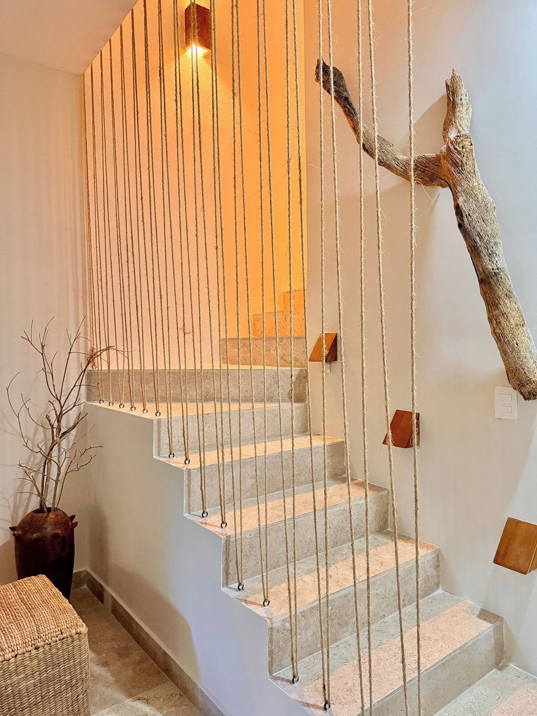 Un escalier avec une barrière en corde, une décoration en forme de branche d’arbre et une plante en pot.