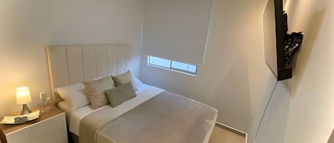 A bedroom with a bed, bedside table, lamp, window, and wall-mounted TV.
